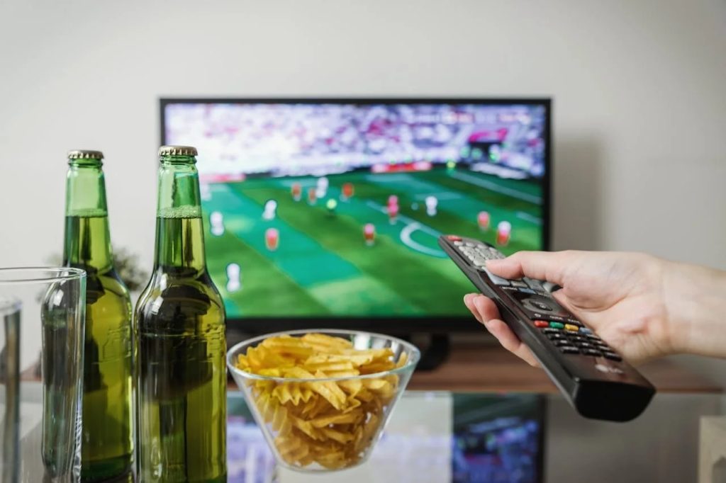Pessoa segurando controle remoto em frente à televisão ligada em jogo de futebol, com garrafas de cerveja e tigela de batatas fritas em primeiro plano, ilustrando hábitos que prejudicam o sono na Copa do Mundo.